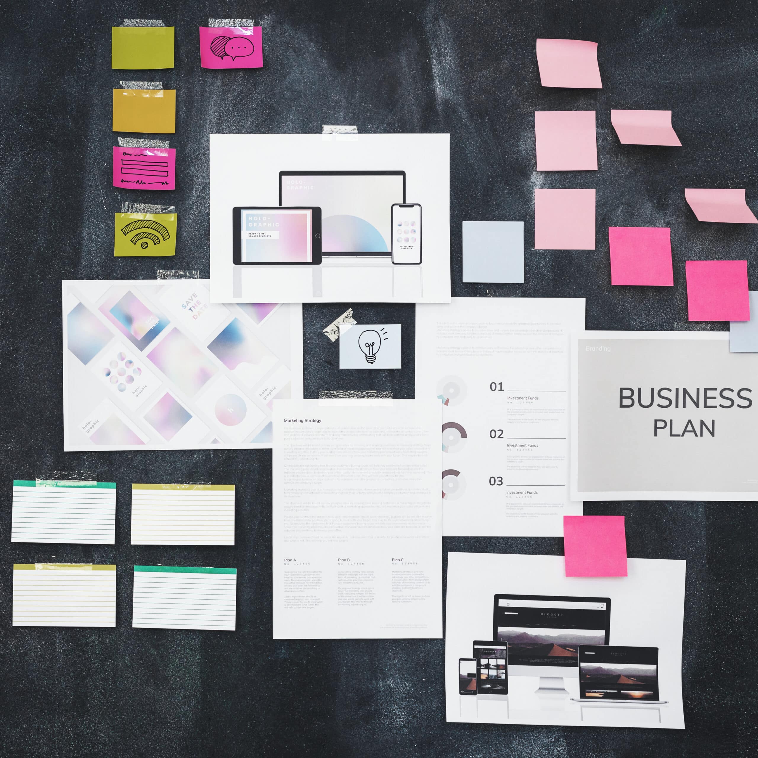 Various documents and sticky notes are arranged on a black surface. The documents include a business plan, marketing strategy, and web designs displayed on digital devices. Pink, yellow, and green sticky notes appear in clusters, some blank and some with sketches.