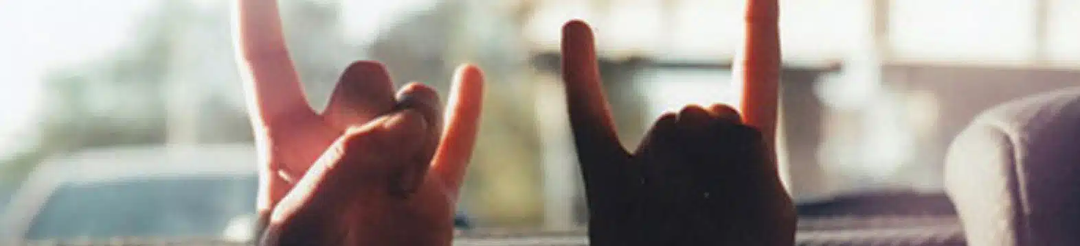 Two hands are shown making the "rock on" or "devil horns" gesture, with the index and pinky fingers extended and the thumb holding down the other fingers. The background is softly blurred, suggesting they are inside a vehicle with outside light streaming in.